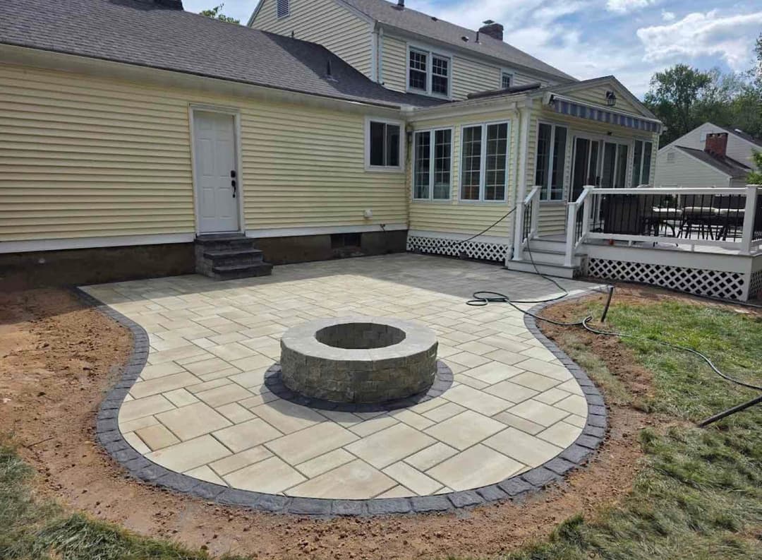 Patio with stone fire pit and paver stones next to a house with large windows and deck.