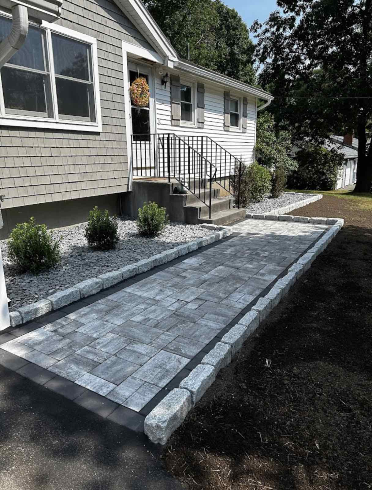 Modern stone walkway leading to a house, bordered with grey stones and landscaped with greenery.