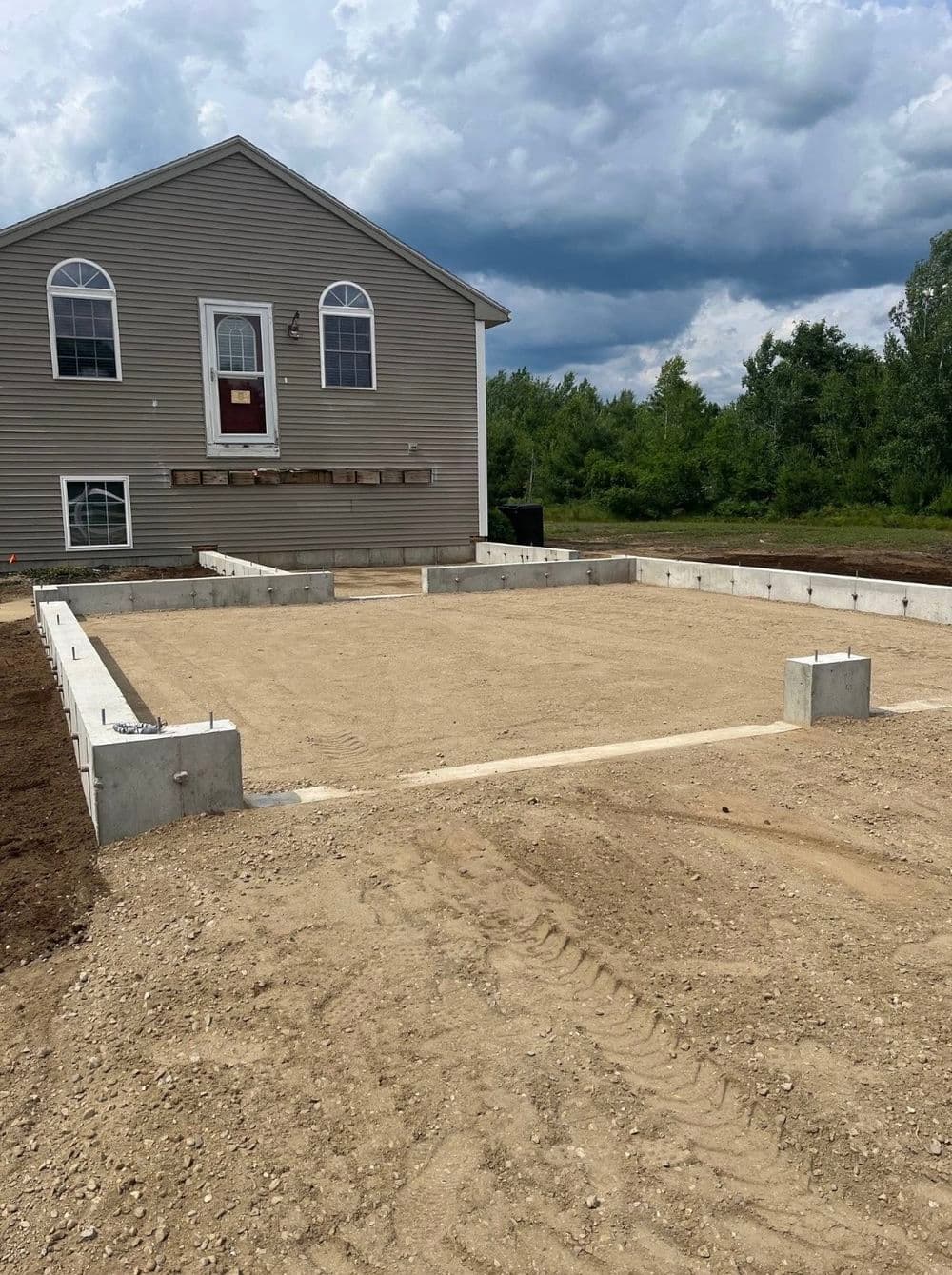 New home foundation on sandy ground with concrete footer and gray house in background.