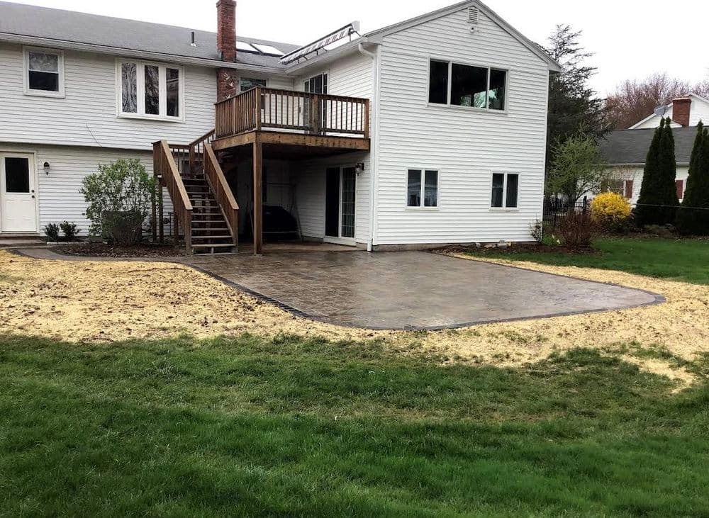 Newly installed concrete patio adjacent to a house with a wooden deck and grassy yard.