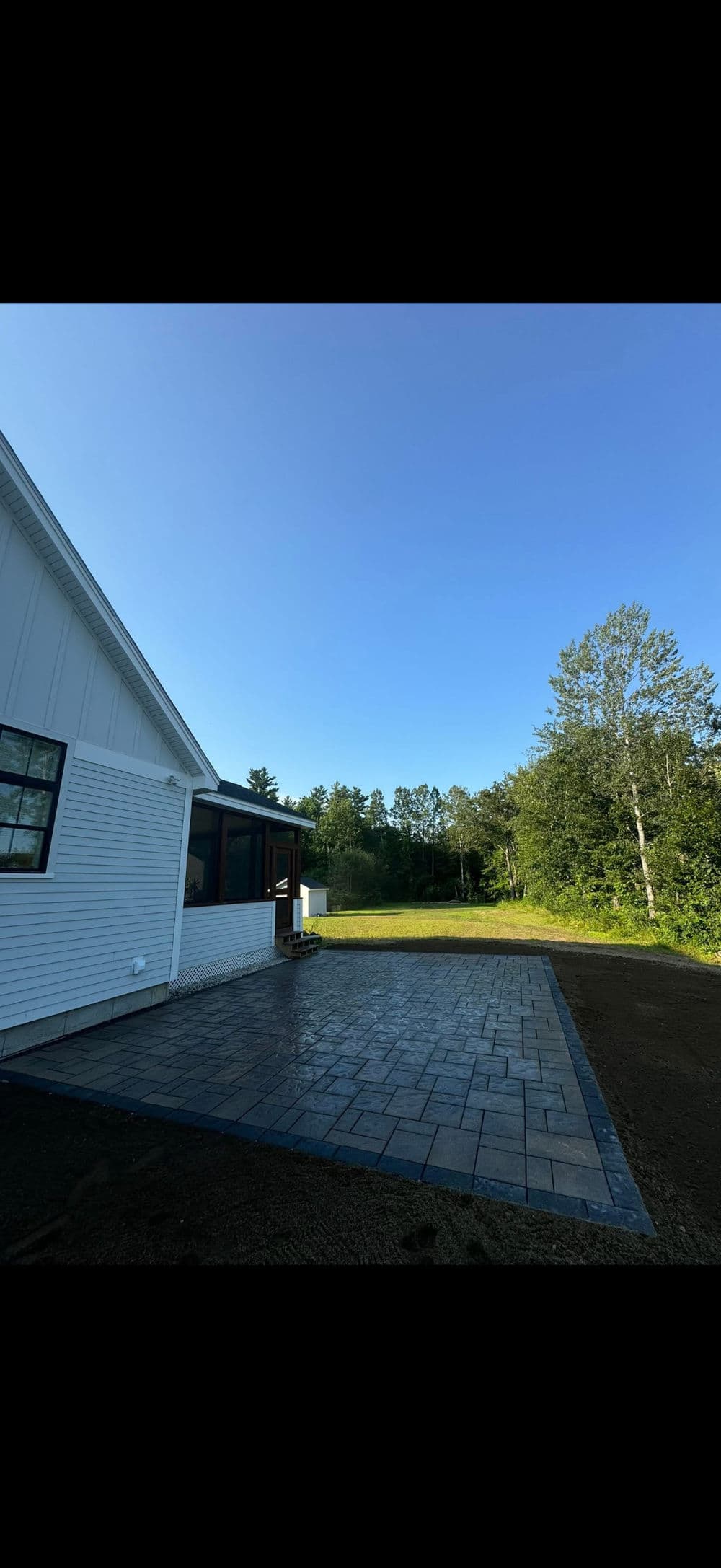 Newly paved patio beside modern white house, surrounded by lush greenery and clear blue sky.